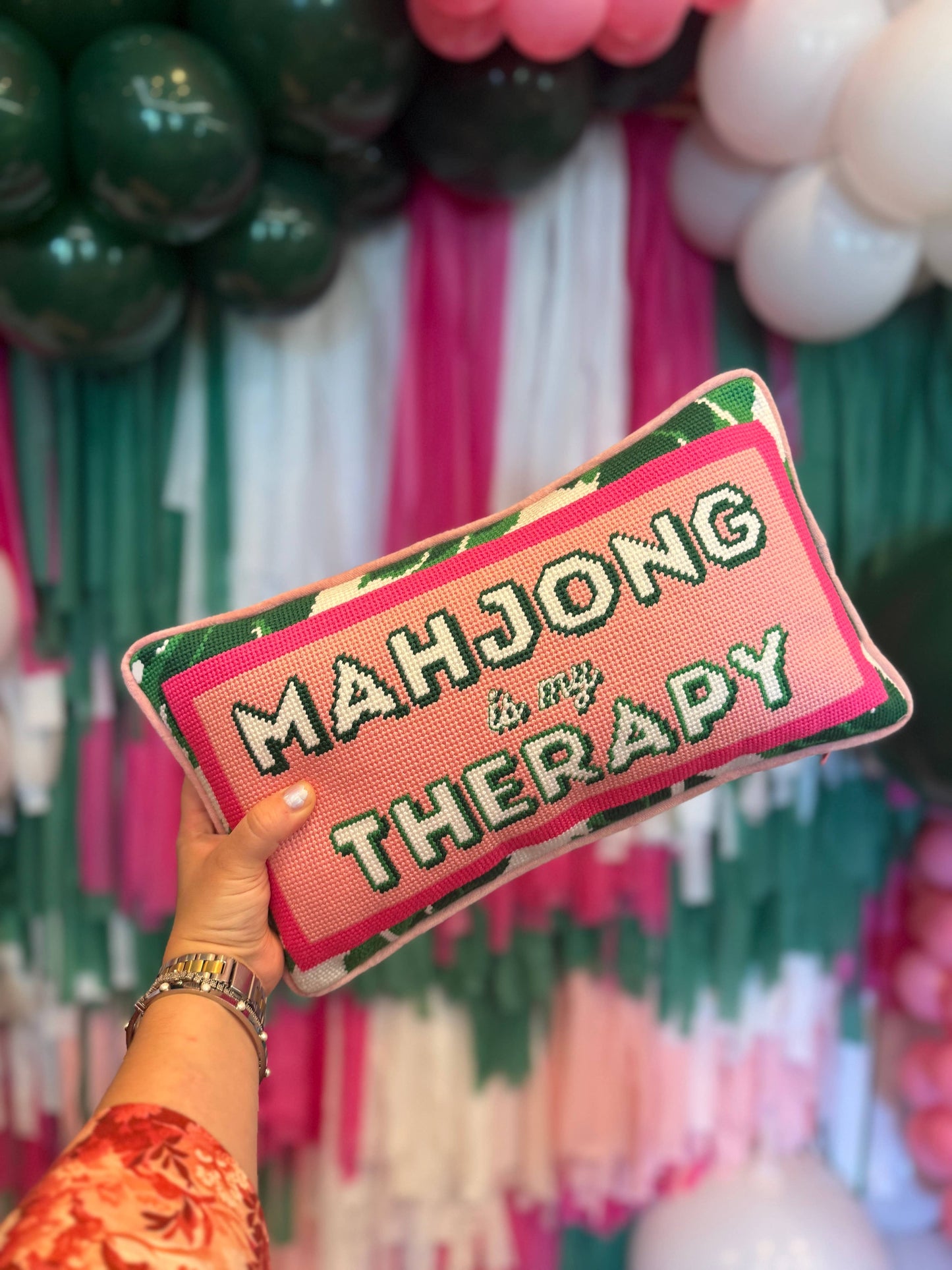 Decorative pillow with 'Mahjong Therapy' text held in front of a colorful balloon arch.