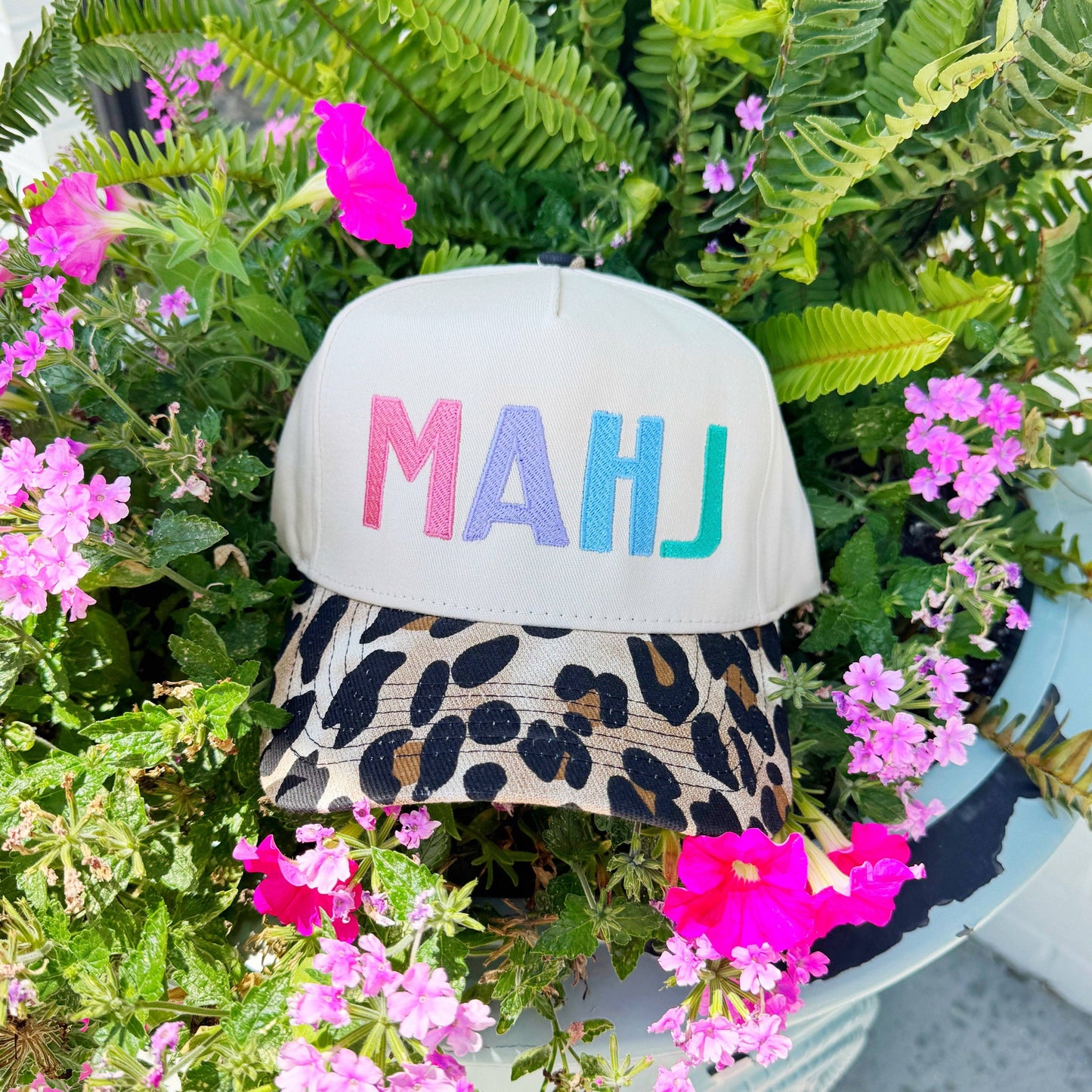 Cap with 'MAHJ' in colorful letters on a plant with pink flowers