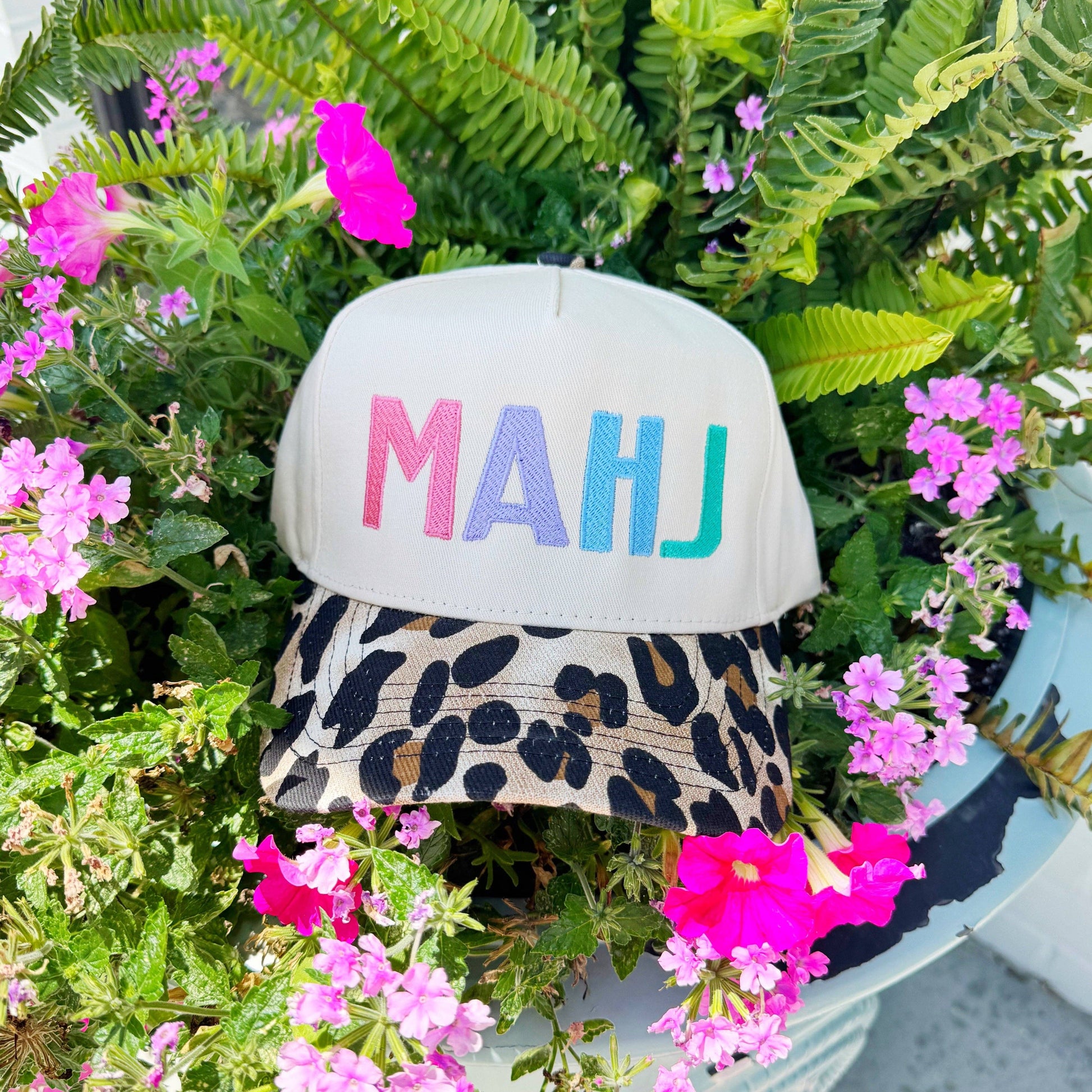 Cap with 'MAHJ' in colorful letters on a plant with pink flowers